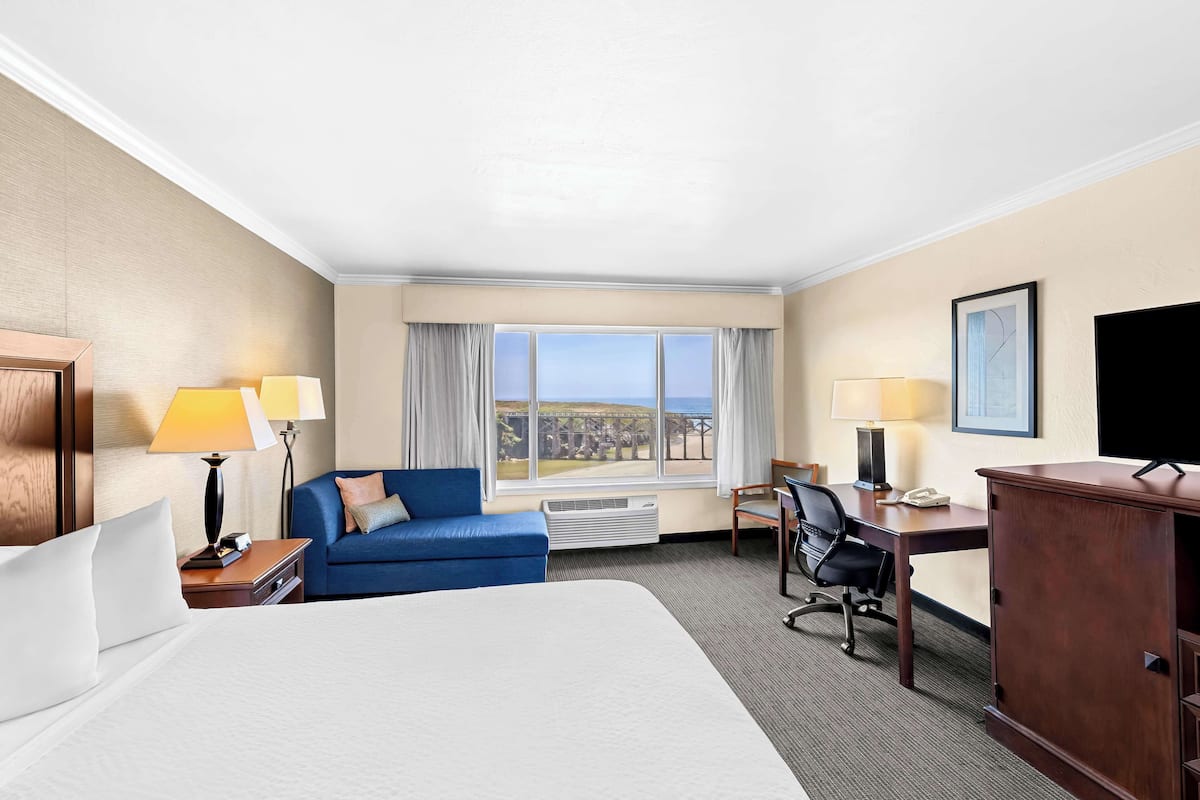 A hotel room with a large bed, a desk with a computer, a chair, a blue sofa, a TV, and a window with a view of the city.