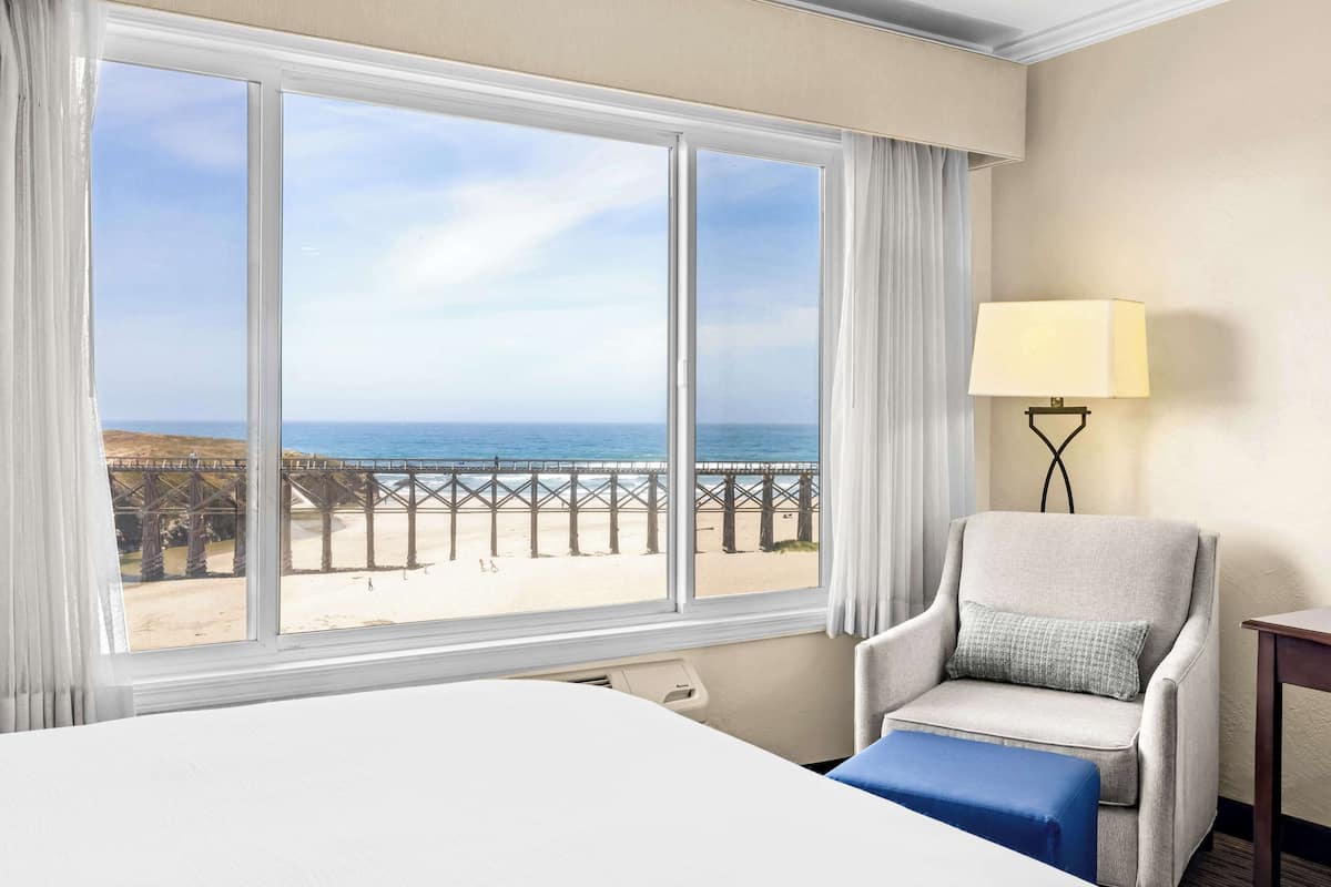A hotel room with a large window overlooking a beach and pier, a bed, a chair with a pillow, and a bedside table with a lamp.