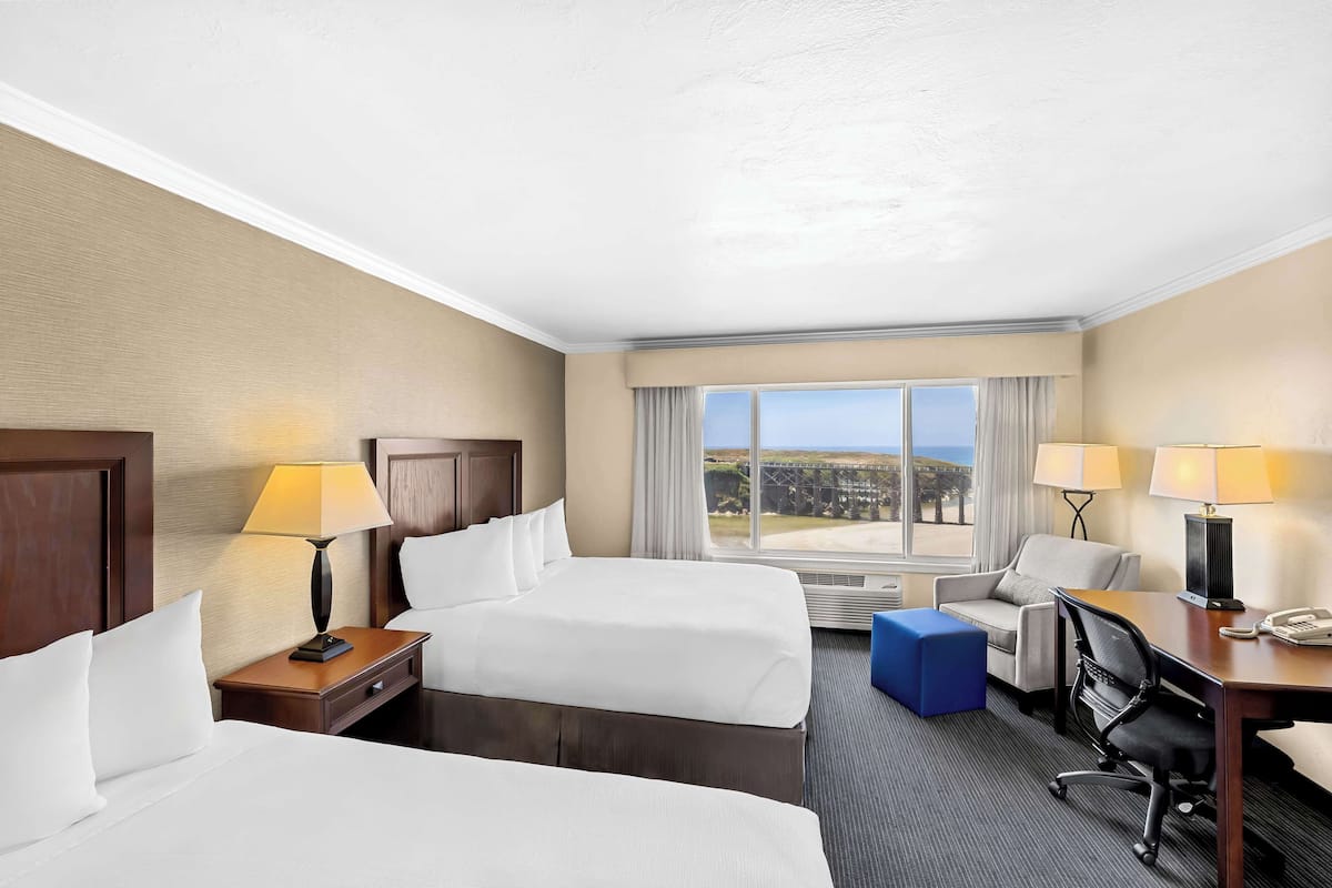 A hotel room with two beds, a desk, a chair, a lamp, and a view of the outdoors.