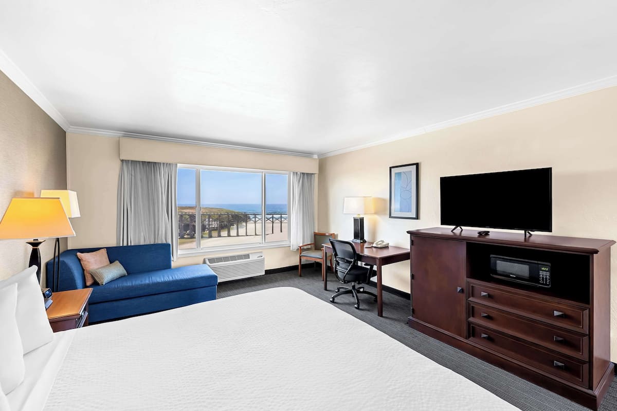 A hotel room with a large bed, a desk with a flat-screen TV, a blue sofa, and a view of the ocean.