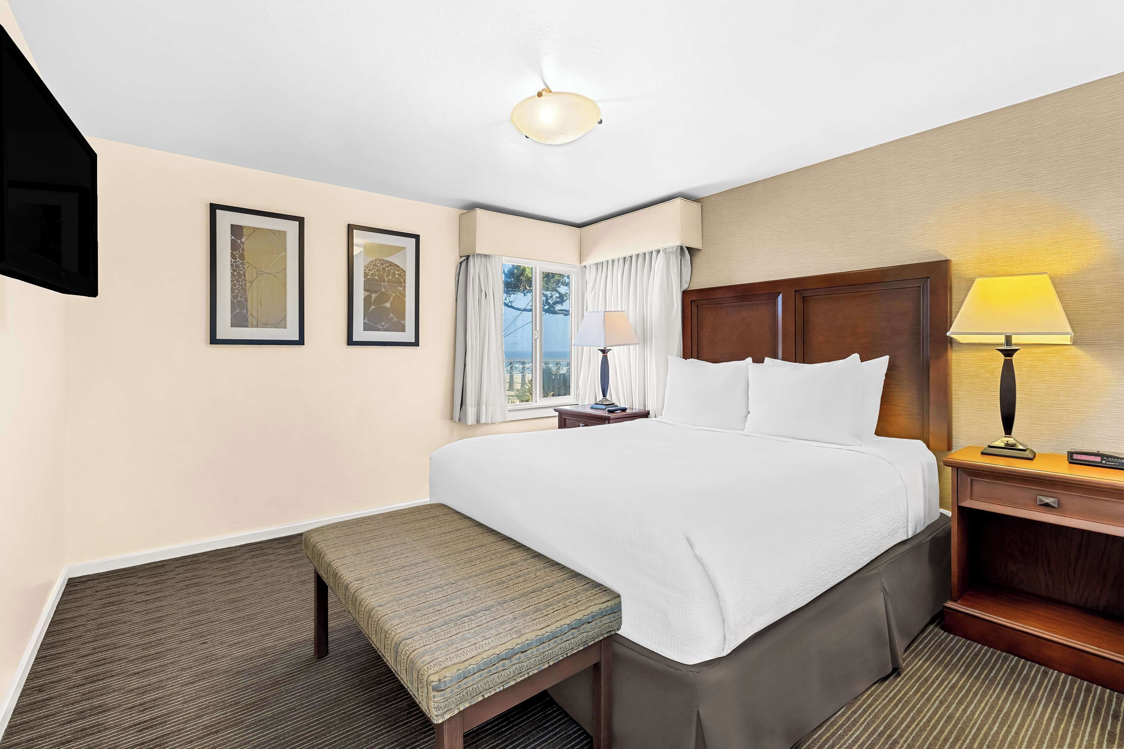 Una habitación de hotel con una cama grande, un banco, una mesita de noche con lámpara y dos cuadros en la pared.