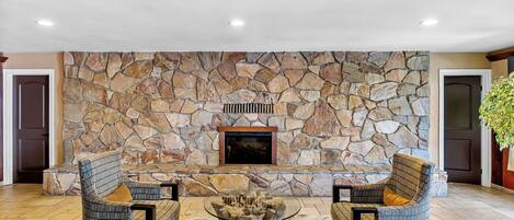 A stone fireplace with two wicker chairs and a glass coffee table in a well-lit room with tiled flooring.