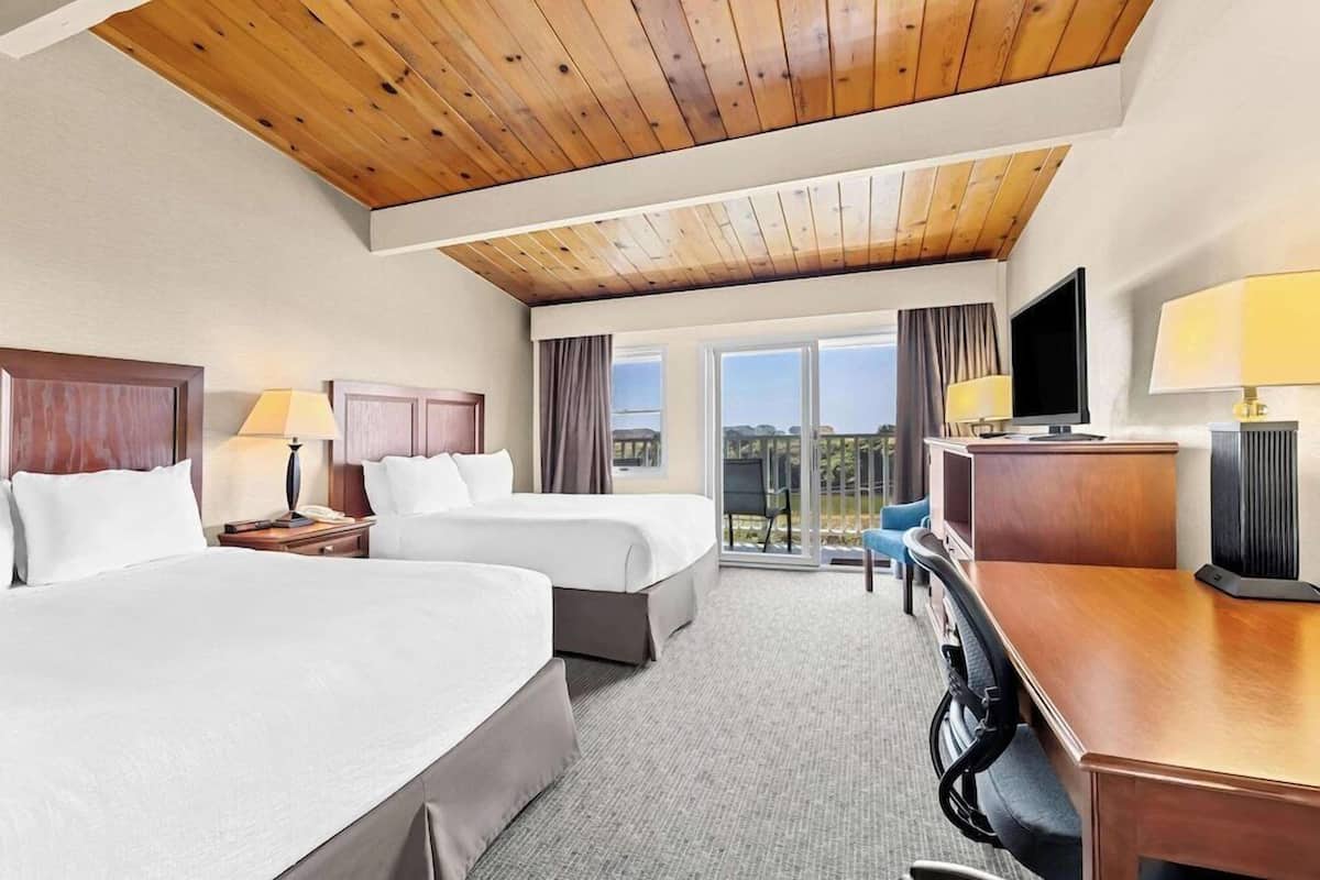 A hotel room with two beds, a desk, a TV, and a view of the outdoors.