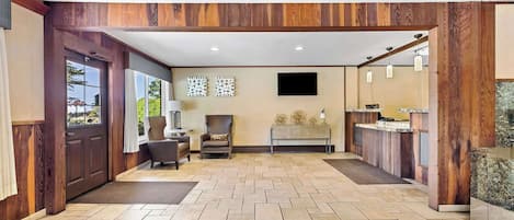 A spacious lobby with a tiled floor, wooden paneling, a reception desk, seating area, and a flat-screen TV mounted on the wall.