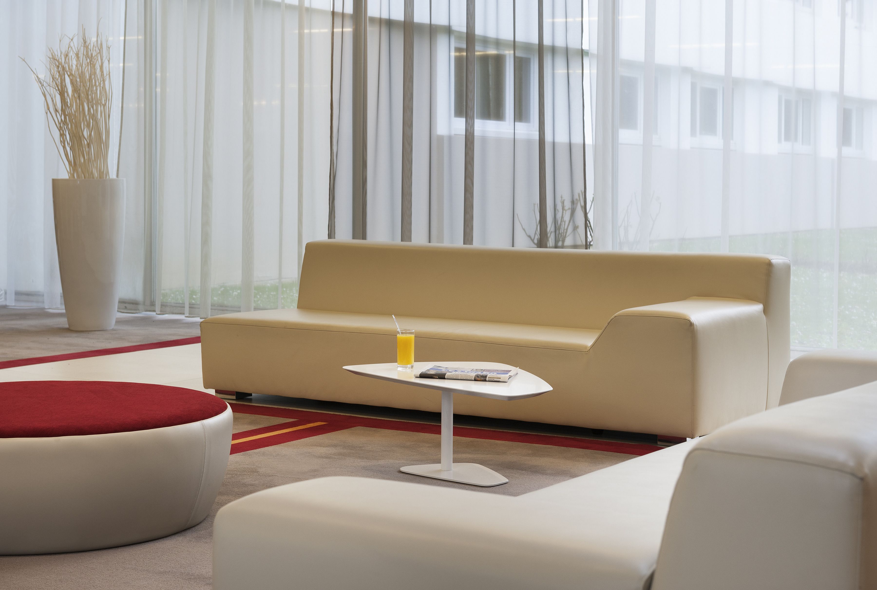 Un salon moderne avec un canapé beige, une petite table blanche sur laquelle se trouve une boisson, et un pouf rouge.