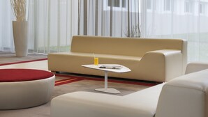 A modern living room with a beige sofa, a small white table with a drink, and a red ottoman. - Mercure Paris Le Bourget (Le Blanc-Mesnil)