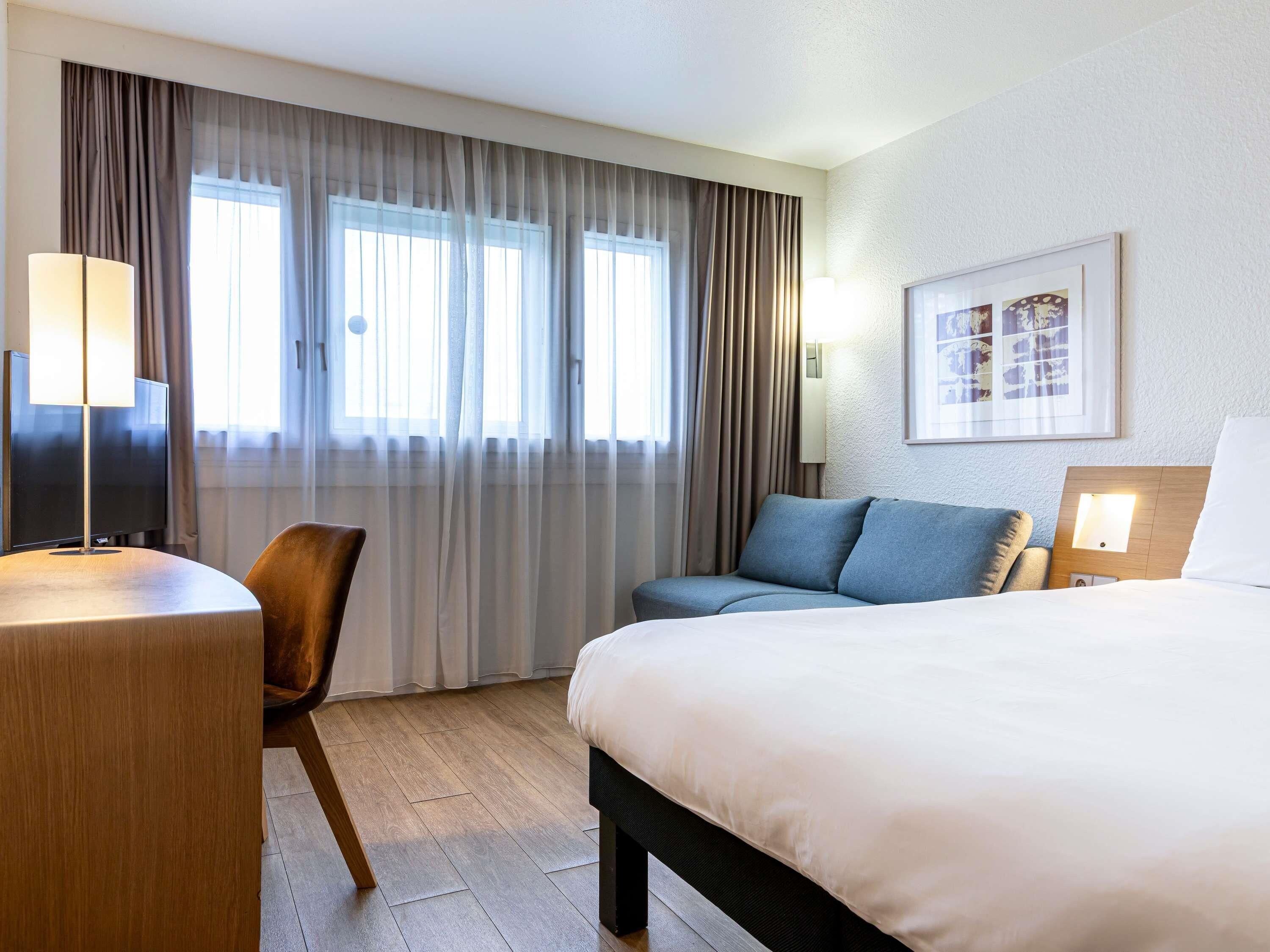 Una habitación de hotel con cama, escritorio, silla, sofá y ventana con cortinas.