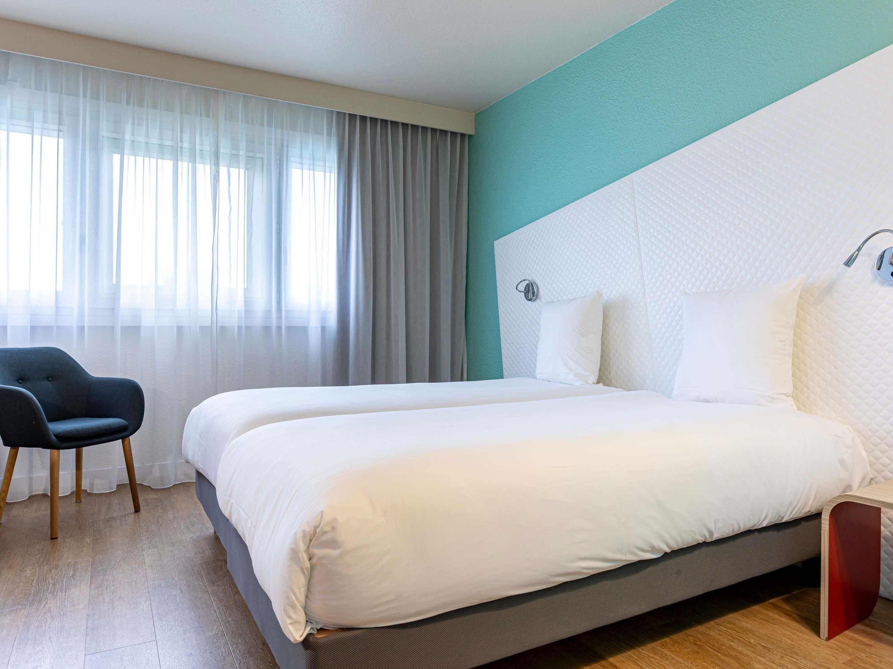 Een hotelkamer met twee bedden, een stoel en een raam met dunne gordijnen.