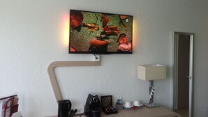 En platt-TV som är monterad på väggen, en lampa, en kaffebryggare och ett litet bord med ett fotoram och dekorativa föremål.