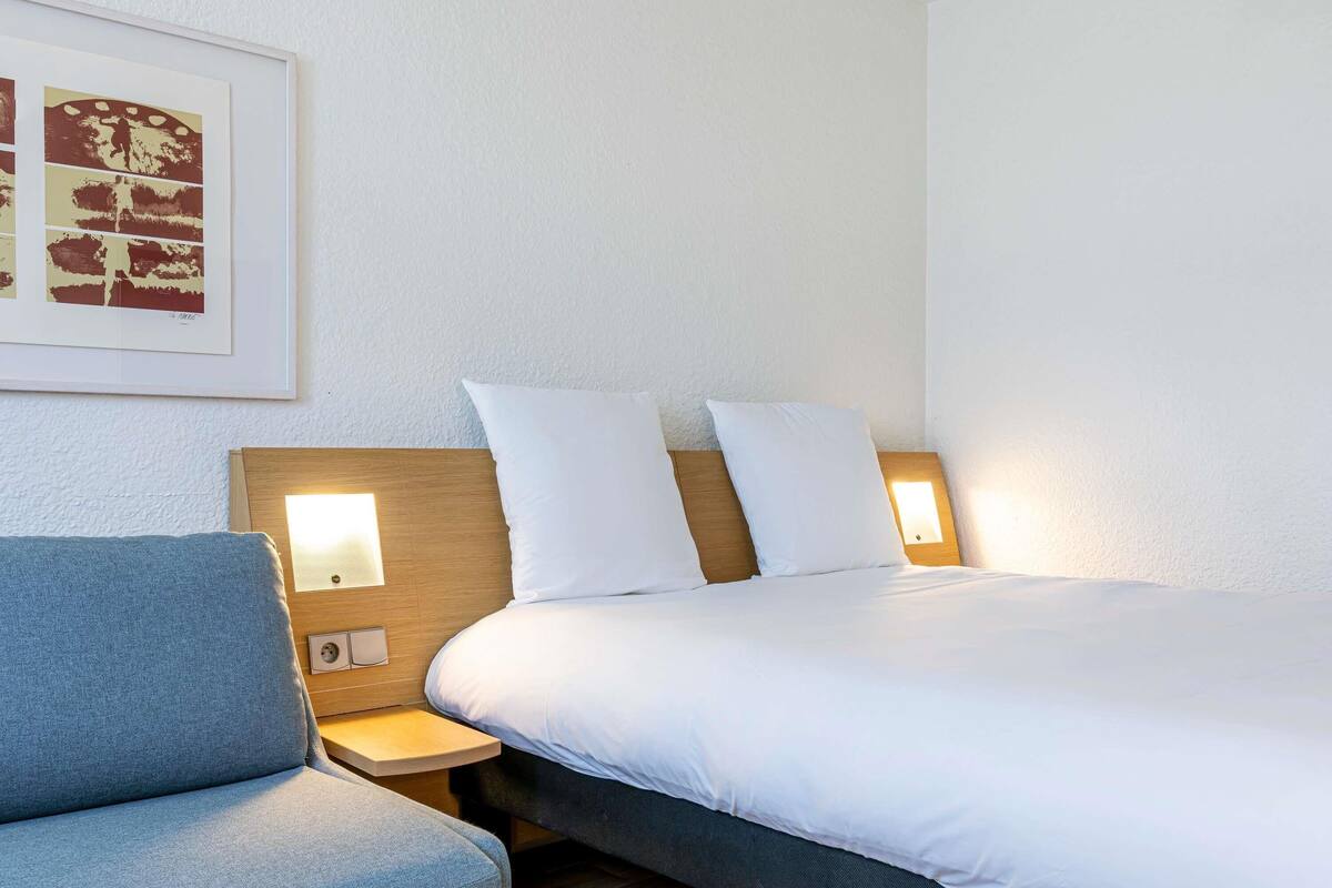 A hotel room with a bed, a nightstand, a chair, and a wall-mounted light fixture.