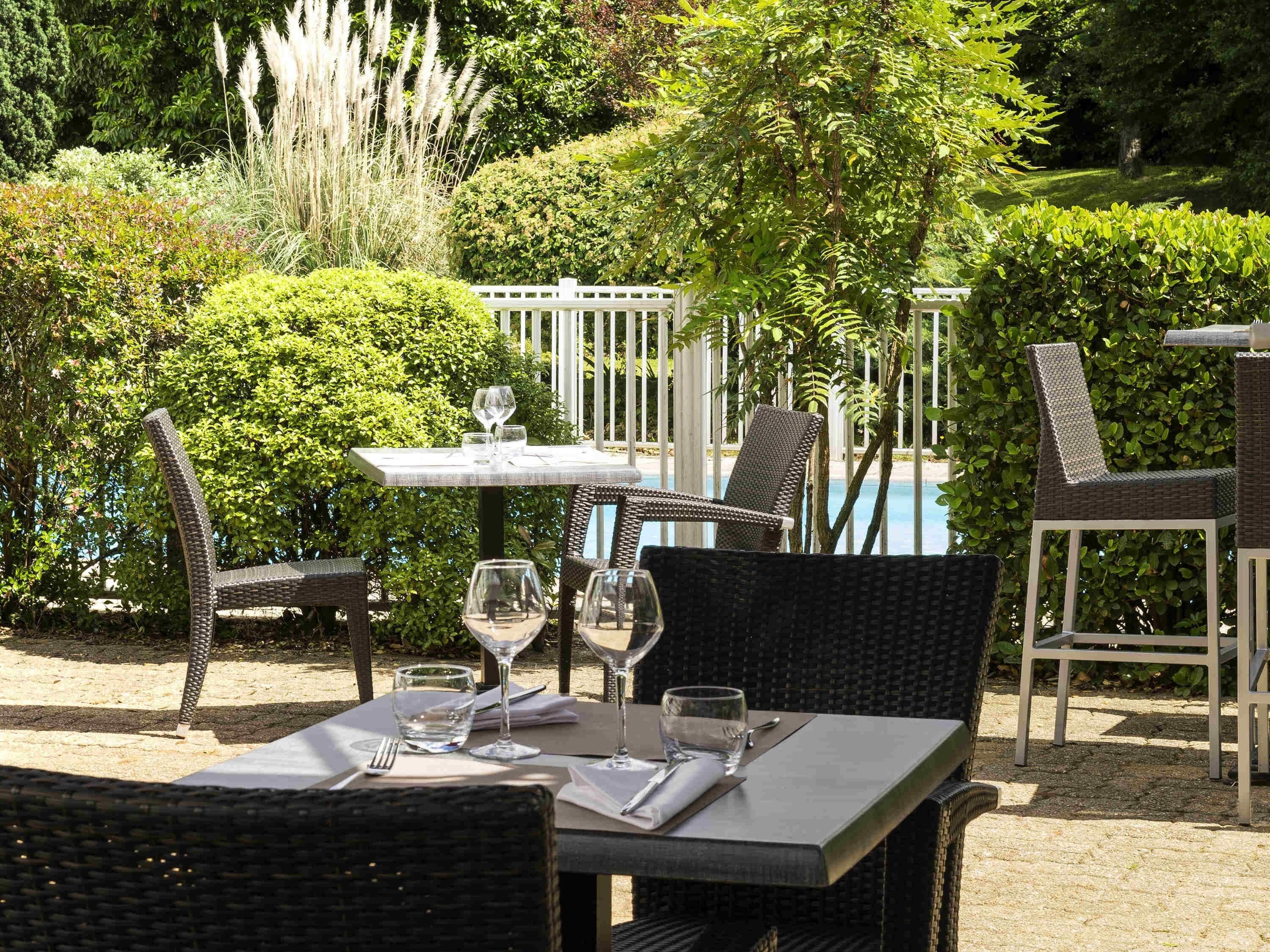 Un espace extérieur aménagé pour les repas, avec des chaises en osier et une table pour deux, entouré d’une végétation luxuriante et avec une piscine en arrière-plan.