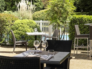 An outdoor dining area with wicker chairs and a table set for two, surrounded by lush greenery and a pool in the background. - Mercure Paris Le Bourget (Le Blanc-Mesnil)