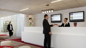 A reception area with a white reception desk, a large screen displaying information, and a vase with flowers. - Mercure Paris Le Bourget (Le Blanc-Mesnil)