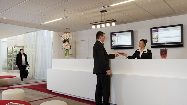 Ett välkomstområde med en vit receptionsdisk, en stor skärm som visar information och en vas med blommor.