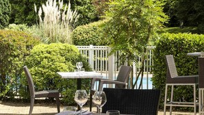 An outdoor dining area with wicker chairs and a table set for two, surrounded by lush greenery and a pool in the background. - Mercure Paris Le Bourget (Le Blanc-Mesnil)