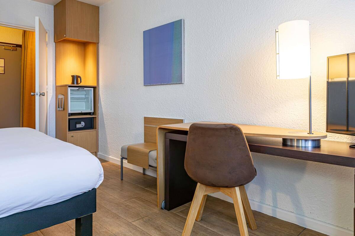 A hotel room with a bed, a desk, a chair, a television, and a wall-mounted lamp.