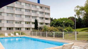 En swimmingpool med solstolar och ett vitt staket, omgiven av grönska och en byggnad med flera våningar.