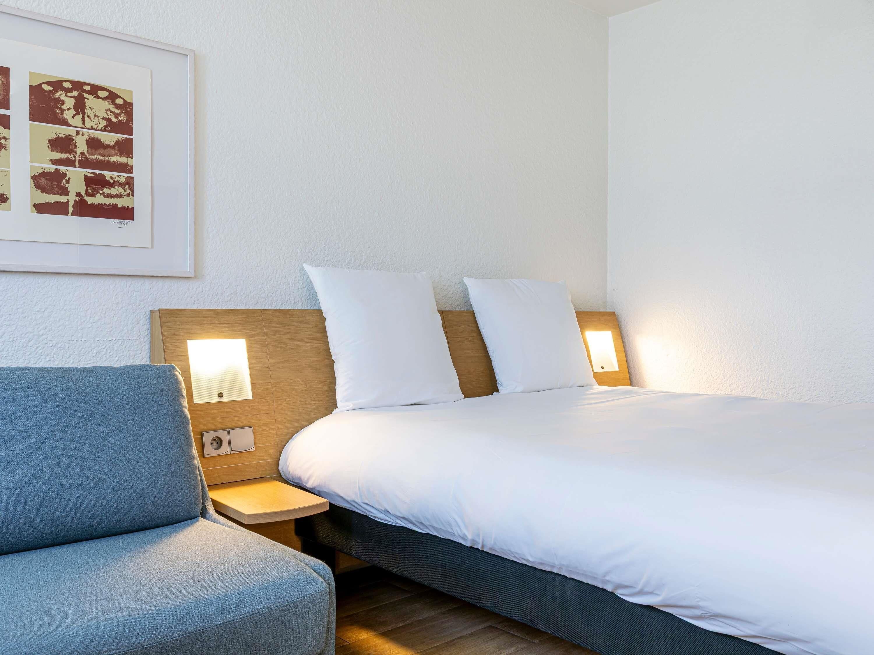 A hotel room with a bed, a nightstand, a chair, and a wall-mounted light fixture.