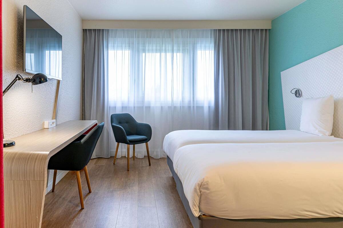 A hotel room with a bed, a desk, a chair, a lamp, and a window with curtains.