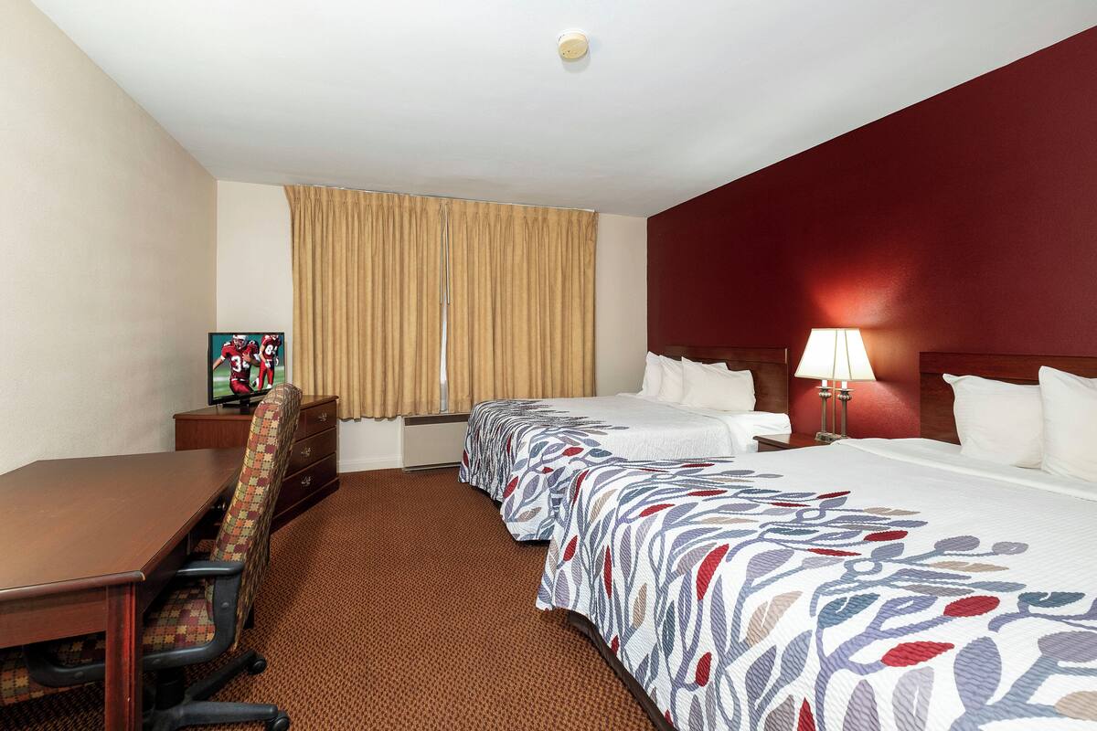 A hotel room with a wooden desk, two beds with floral bedspreads, a television, a window with curtains, and a nightstand with a lamp.