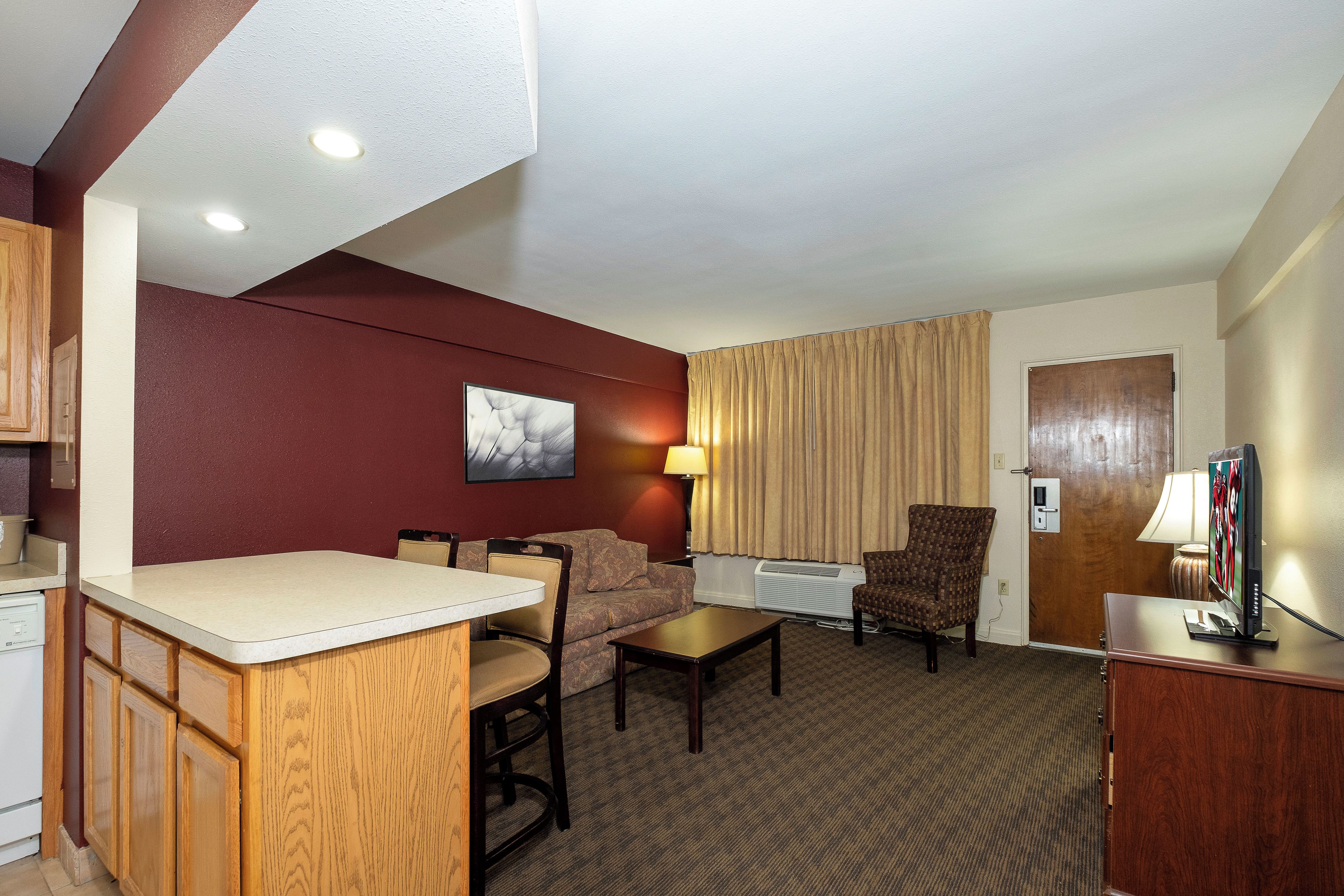 A hotel room with a kitchenette, a dining area, a living area with a sofa and armchair, a television, and a window with curtains.
