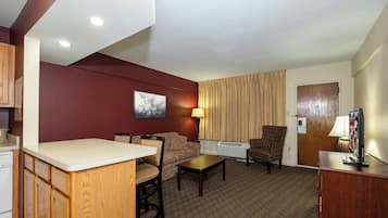 A hotel room with a kitchenette, a dining area, a living area with a sofa and armchair, a television, and a window with curtains.