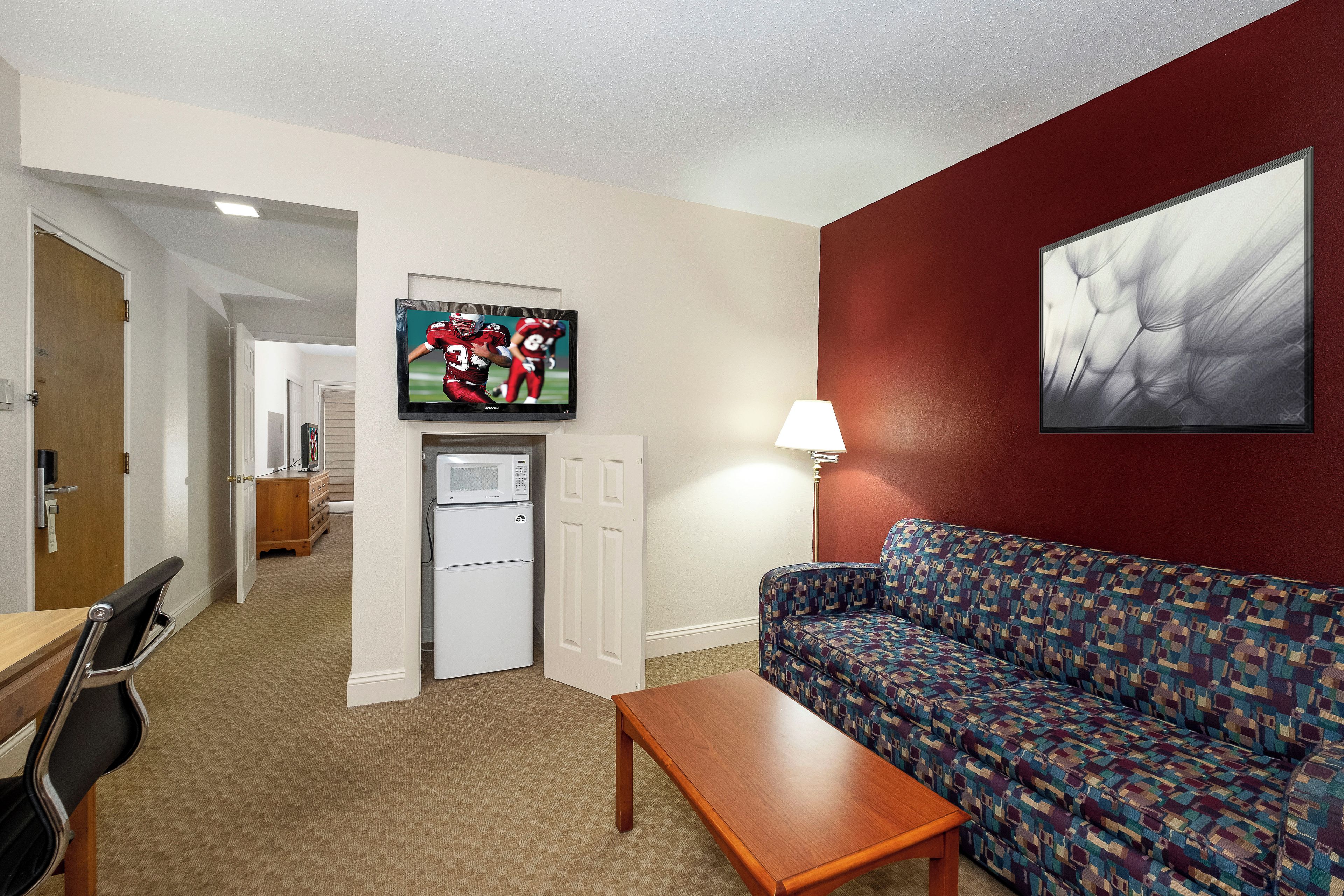 A living room with a sofa, a coffee table, a television mounted on the wall, a refrigerator, and a door leading to another room.
