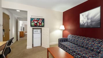 A living room with a sofa, a coffee table, a television mounted on the wall, a refrigerator, and a door leading to another room.