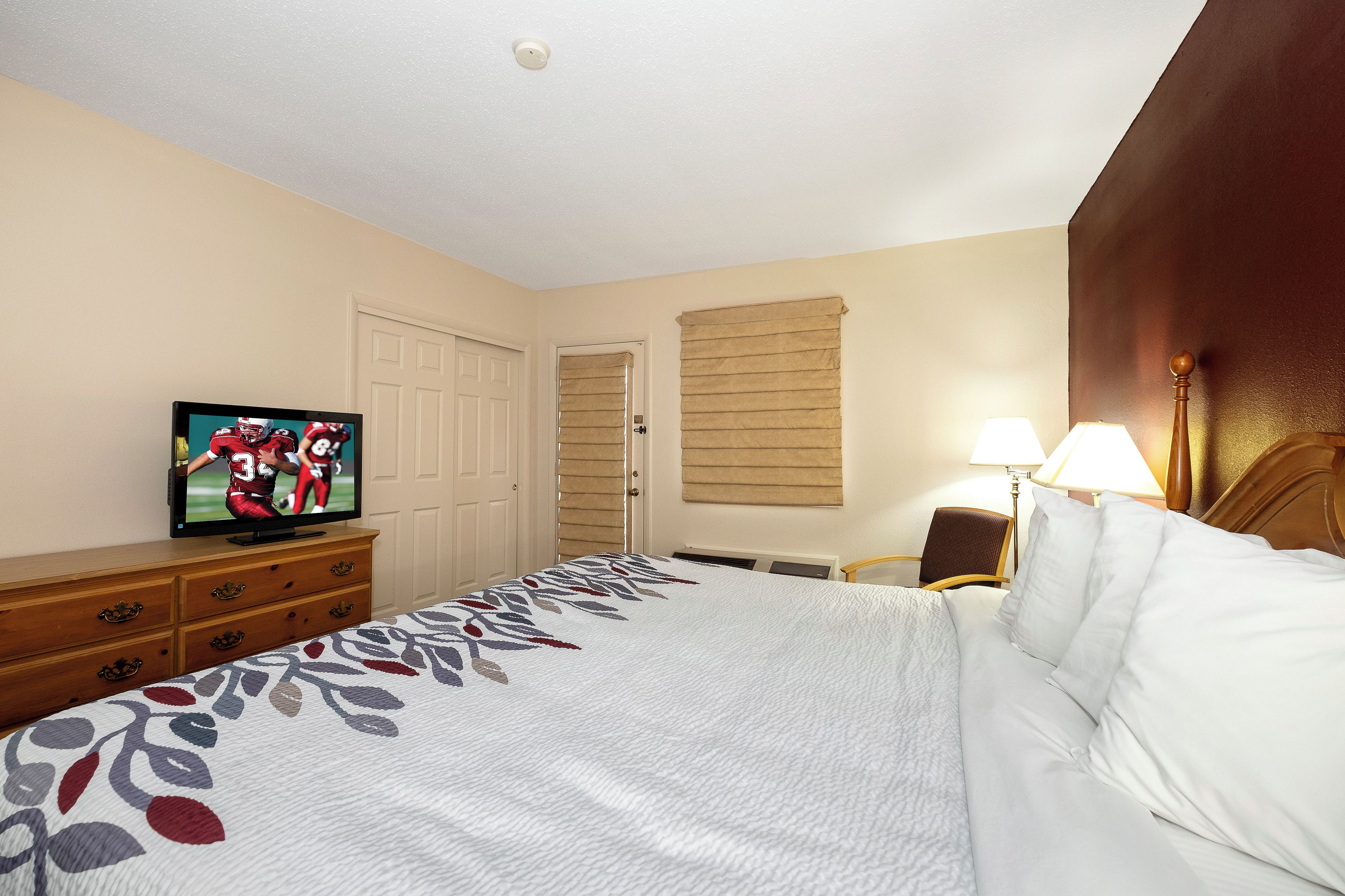 Habitación de hotel con una cama, un televisor sobre un mueble de madera, una puerta, una ventana con persianas y una lámpara.