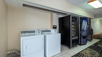 A hotel laundry room with two front-loading washing machines, a vending machine, and a refrigerator.