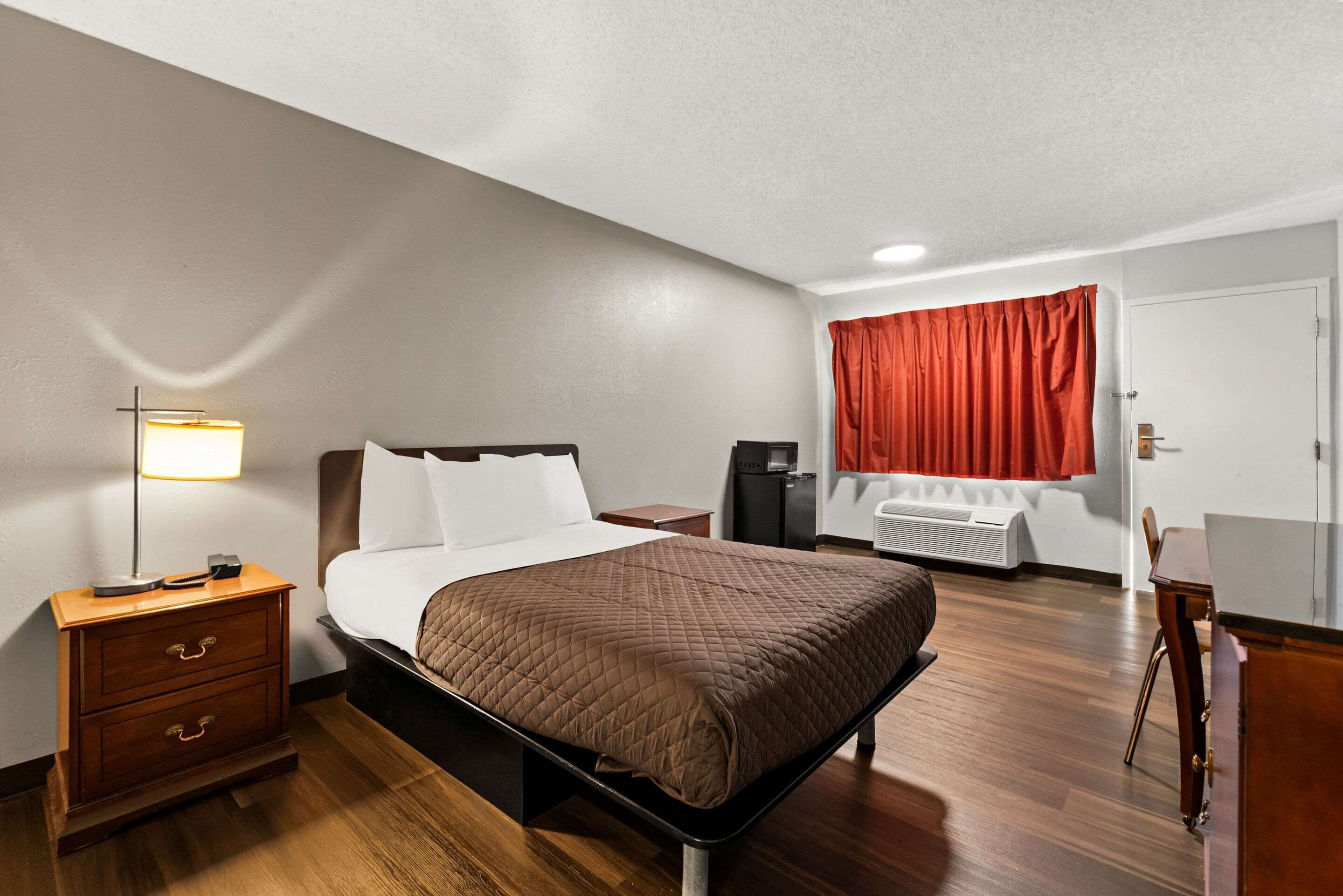 A hotel room with a bed, bedside table, lamp, and a desk with a chair.