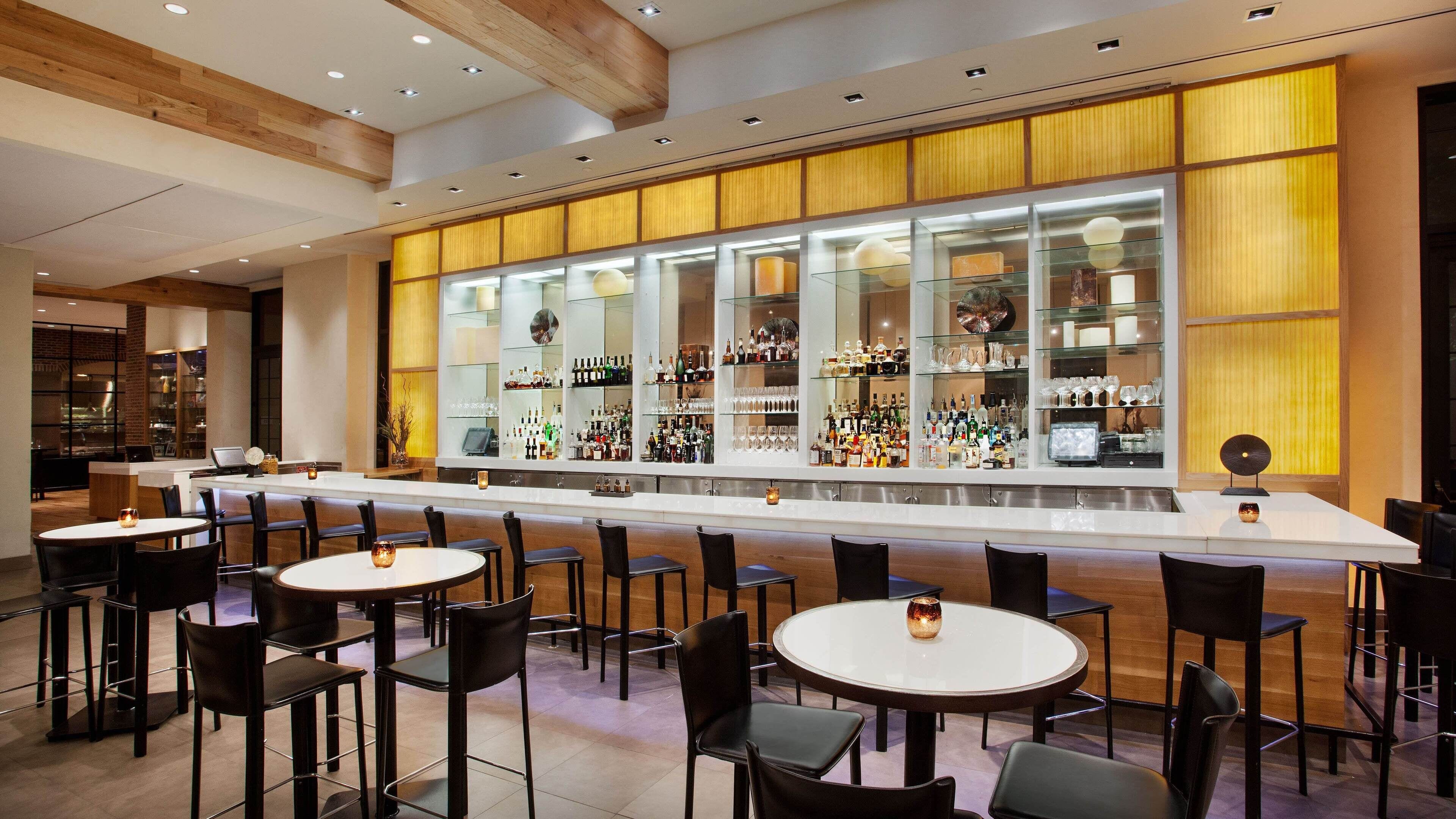 A modern bar area with a long counter, multiple seating options, and a variety of bottles and glasses displayed in glass cases.