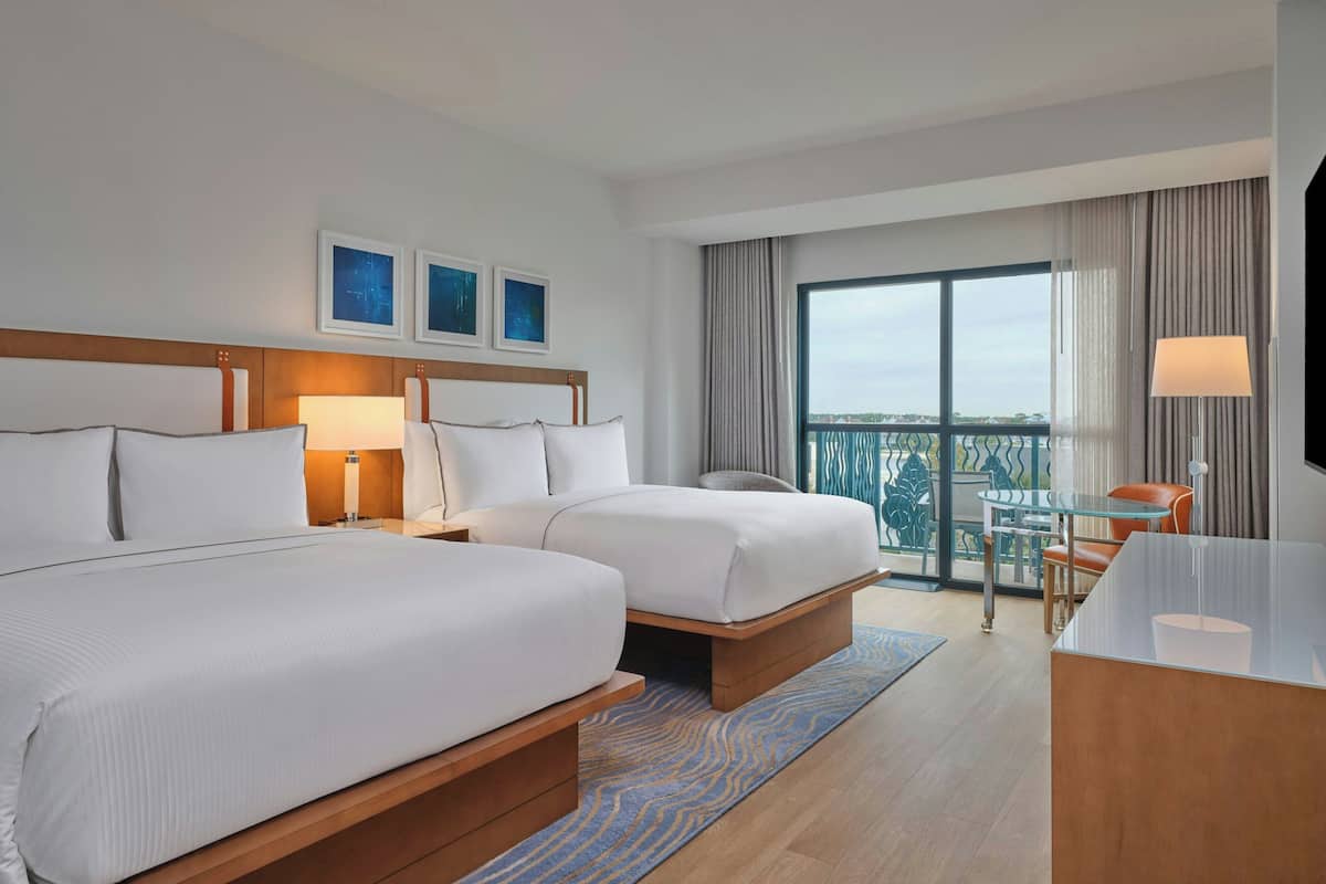 A hotel room with two beds, a desk, a chair, a TV, and a balcony with a view.