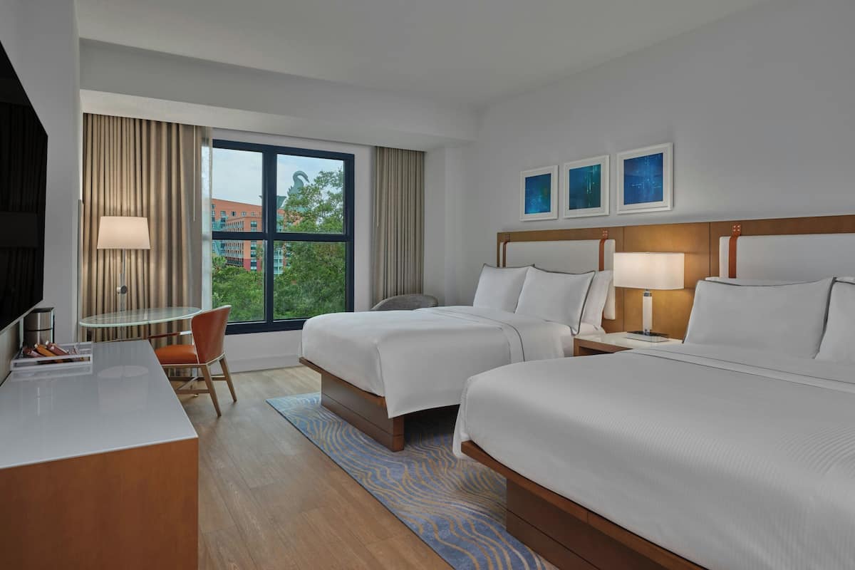 A hotel room with two beds, a desk, a chair, a TV, and a window with a view of buildings and trees.