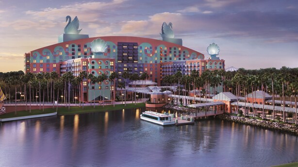 A large, multi-story hotel with a distinctive swan-shaped roof ornament, a waterfront view, and a boat dock. - Walt Disney World Swan (Lake Buena Vista)