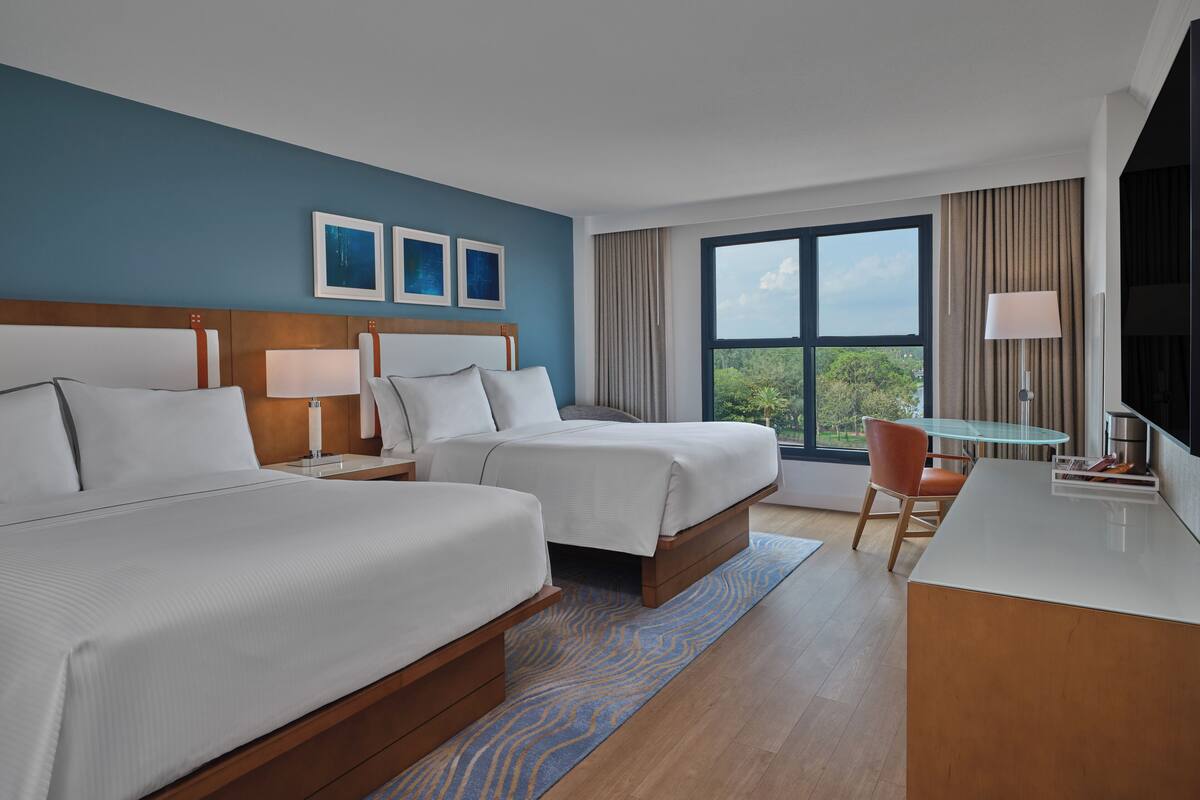 A hotel room with two beds, a desk, a chair, and a large window offering a view of greenery.