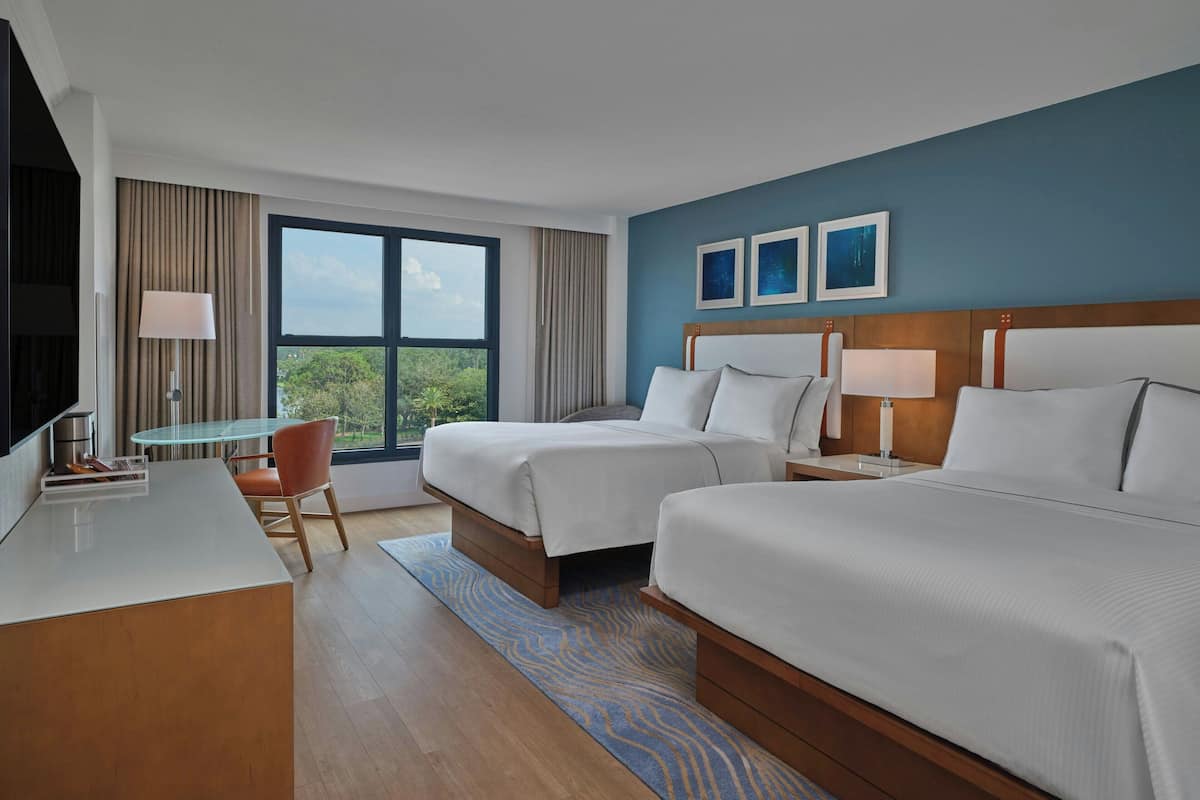 A hotel room with two beds, a desk, a chair, and a large window offering a view of greenery.