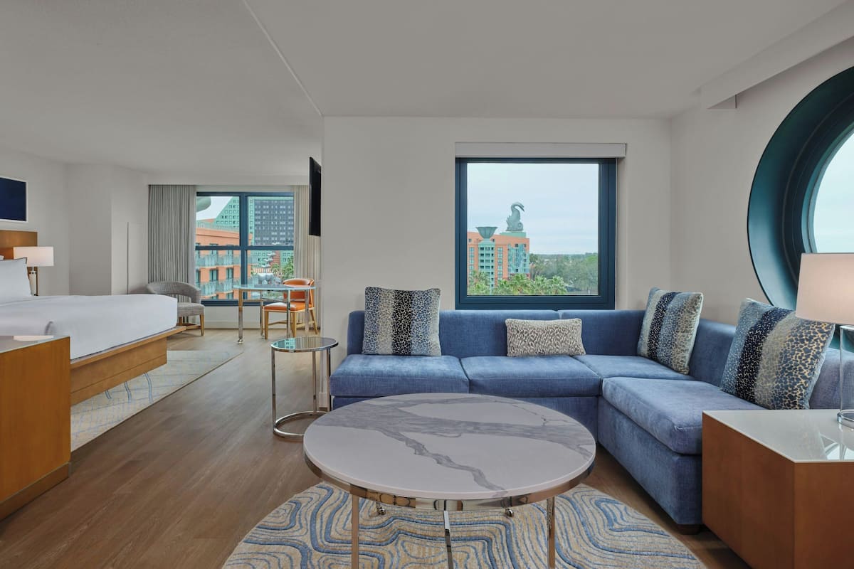 A modern hotel room with a bed, sofa, and round coffee table. There is a view of buildings through large windows.