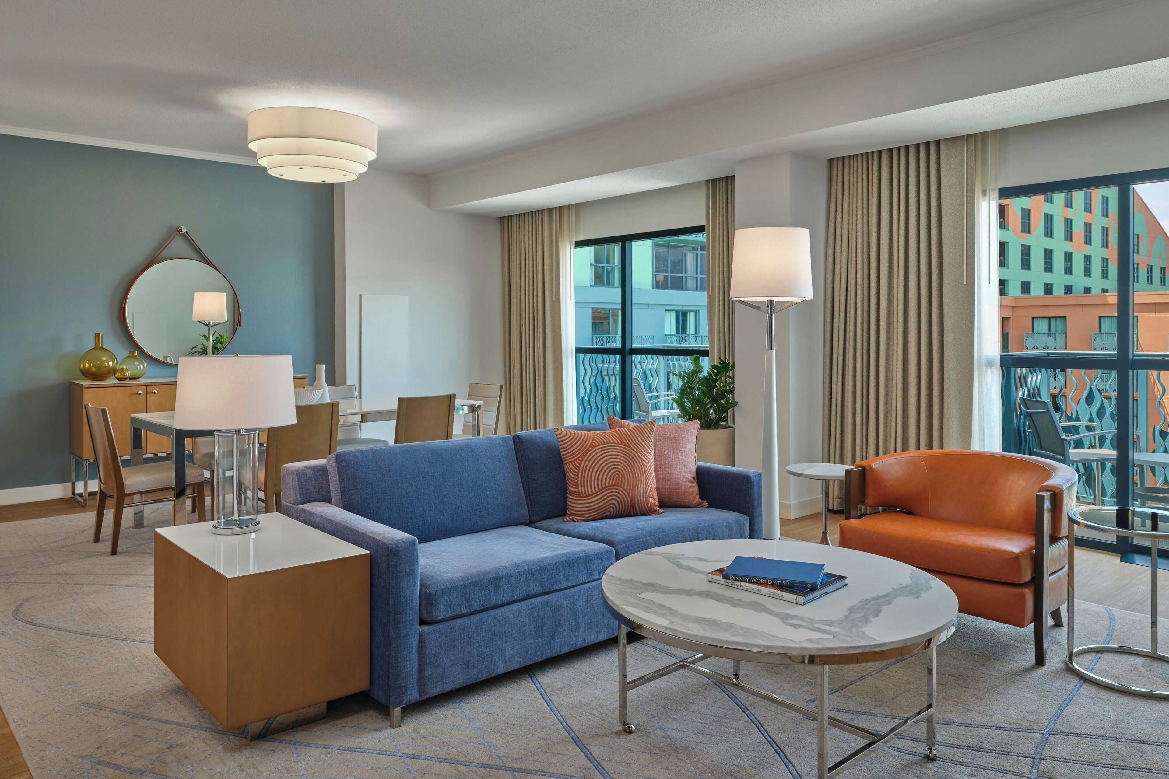 Una habitación de hotel con un sofá azul, un sillón naranja, una mesa de centro redonda y un área de comedor con una mesa y sillas.