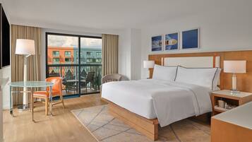 A hotel room with a large bed, a glass table with a chair, a small desk, and a view of buildings through a large window.