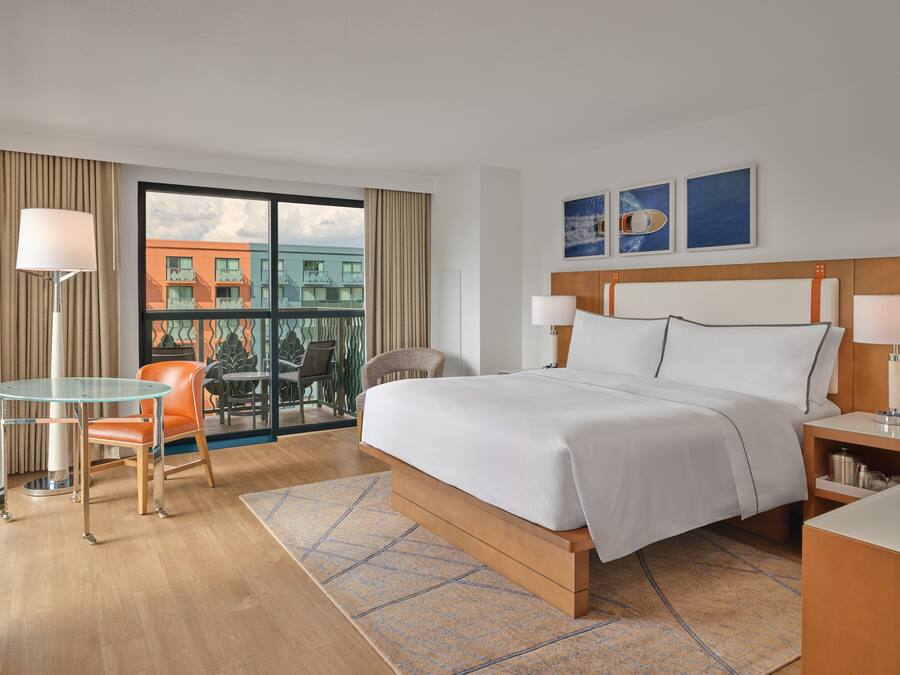 A hotel room with a large bed, a glass table with a chair, a small desk, and a view of buildings through a large window.