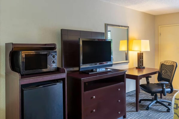 Habitación de hotel con microondas, televisión de pantalla plana, escritorio con lámpara, silla y refrigerador pequeño.