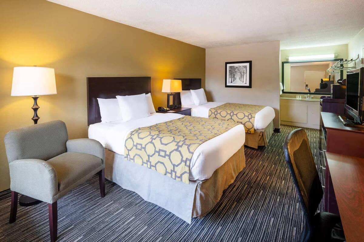 A hotel room with two beds, a desk, a chair, a lamp, a picture on the wall, and a bathroom with a mirror and sink.