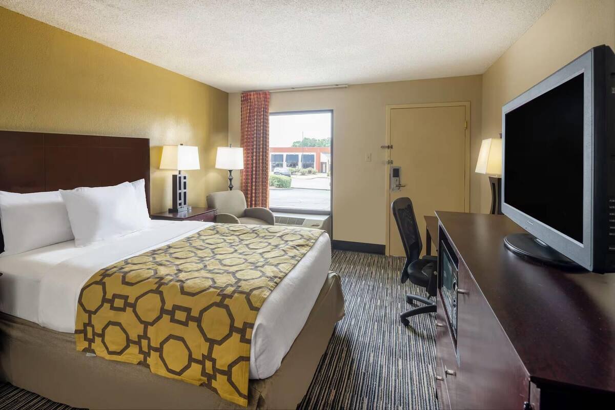 A hotel room with a bed, two bedside lamps, a desk with a flat-screen TV, and a view of the outside.