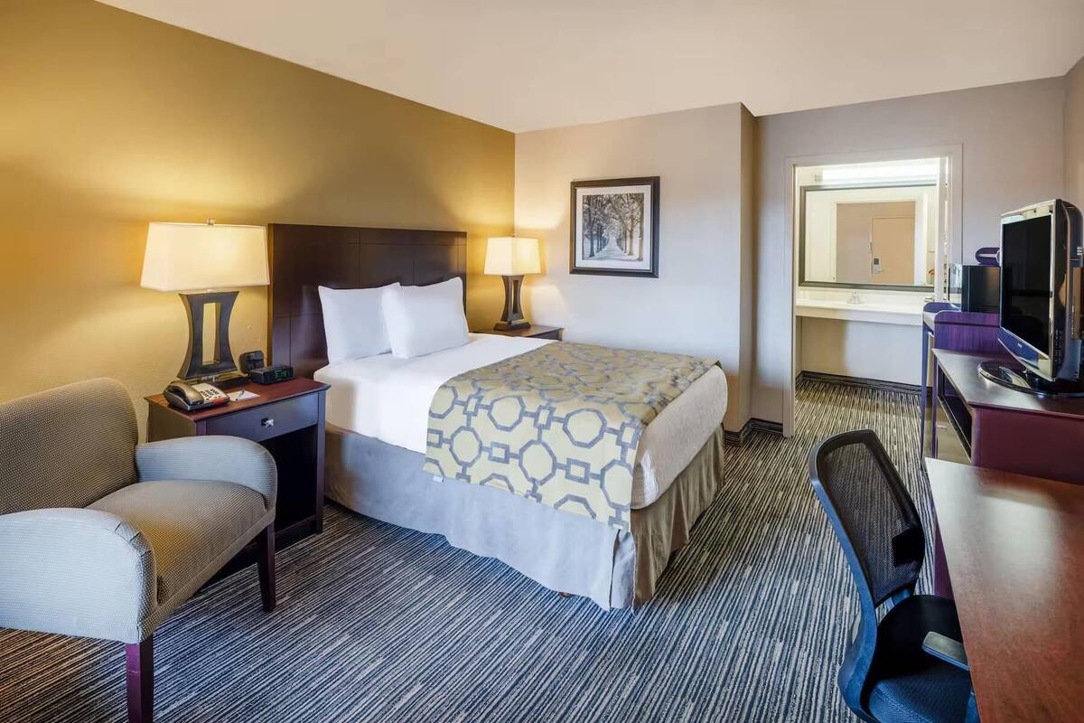 A hotel room with a bed, two bedside lamps, a chair, a desk with a computer, a television, and a mirror.