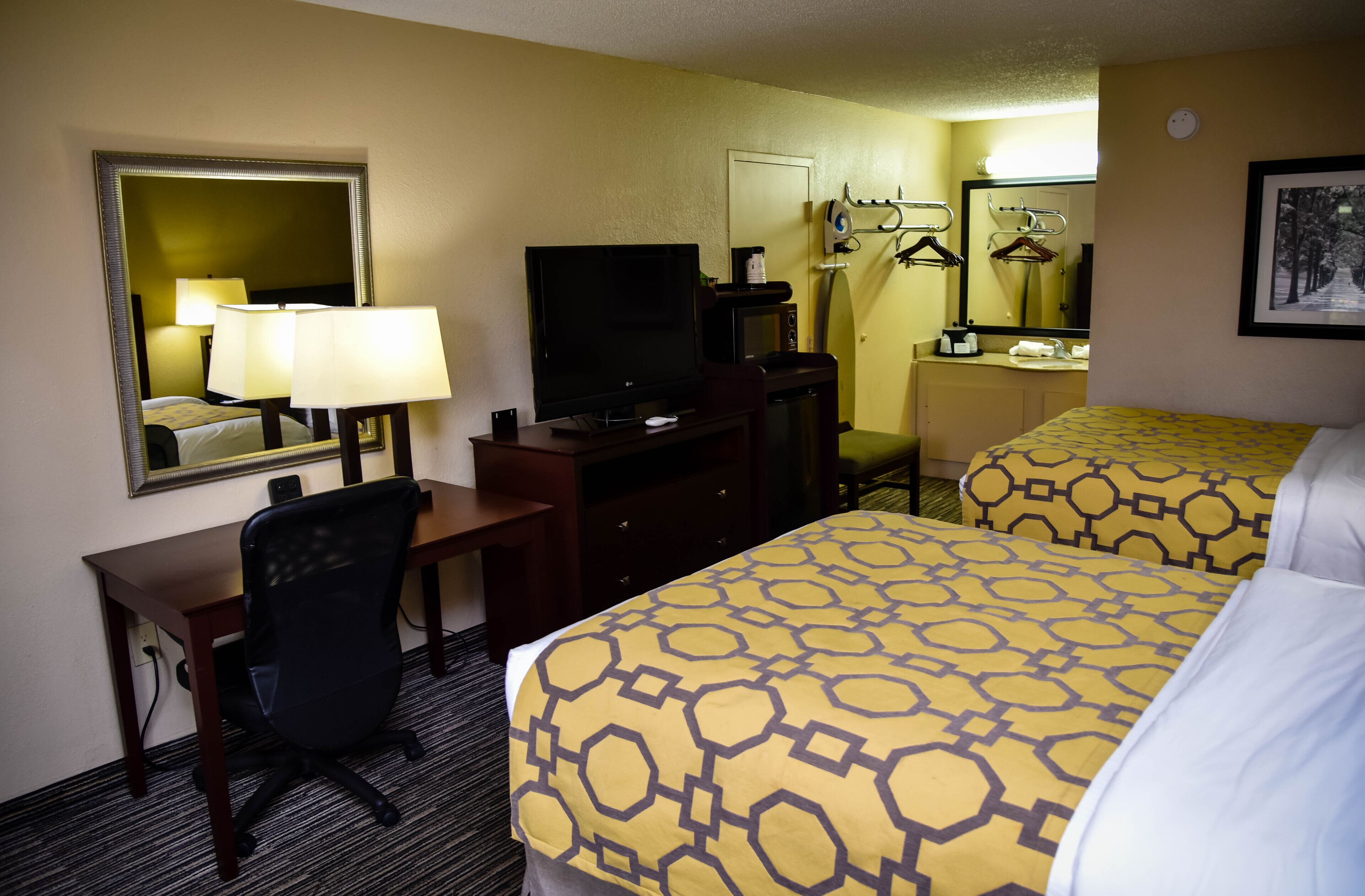 A hotel room with a desk, chair, television, two beds with patterned bedding, a mirror, and a coat rack.