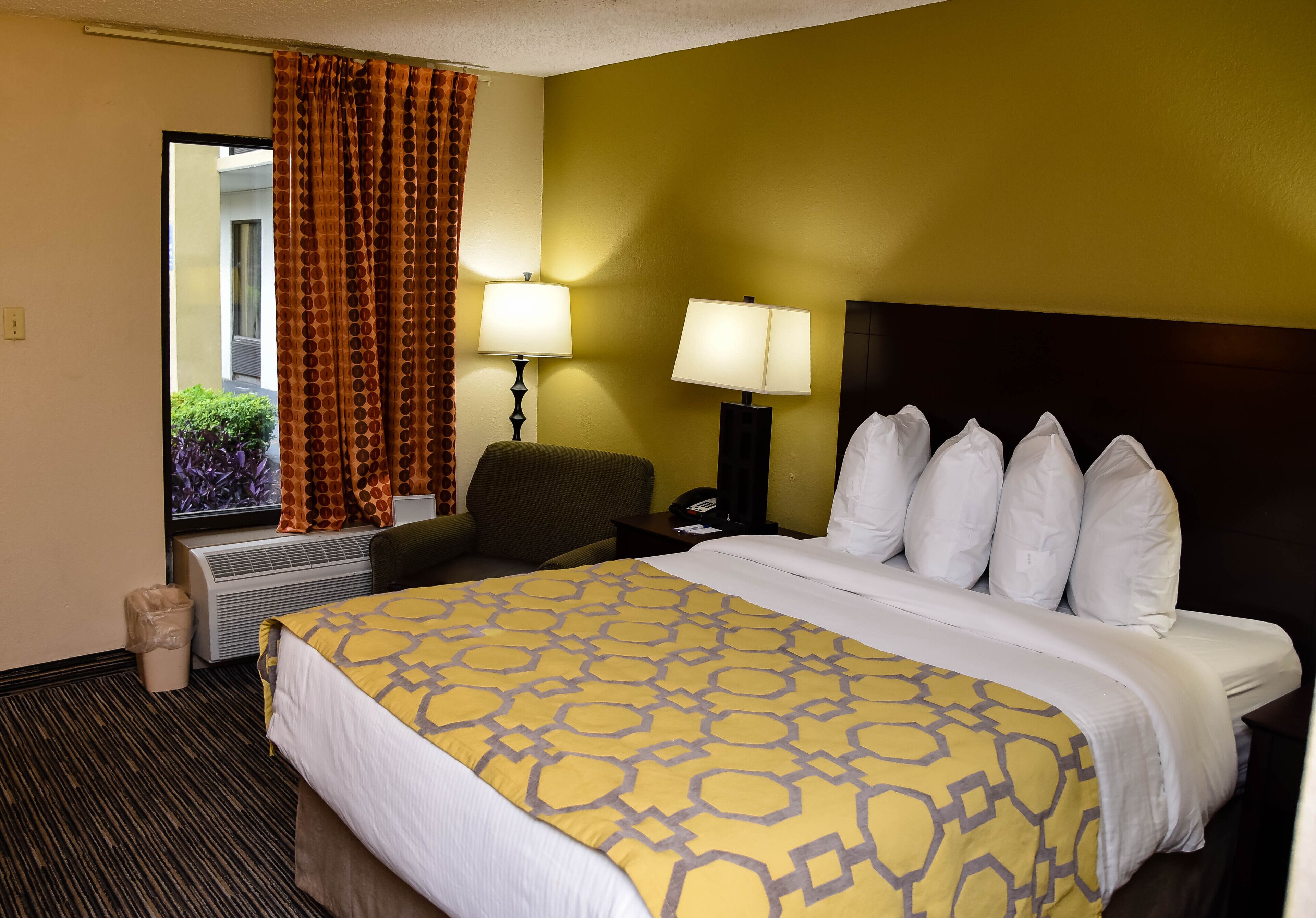 A hotel room with a bed, two bedside lamps, a chair, a desk, and a view of the outside.