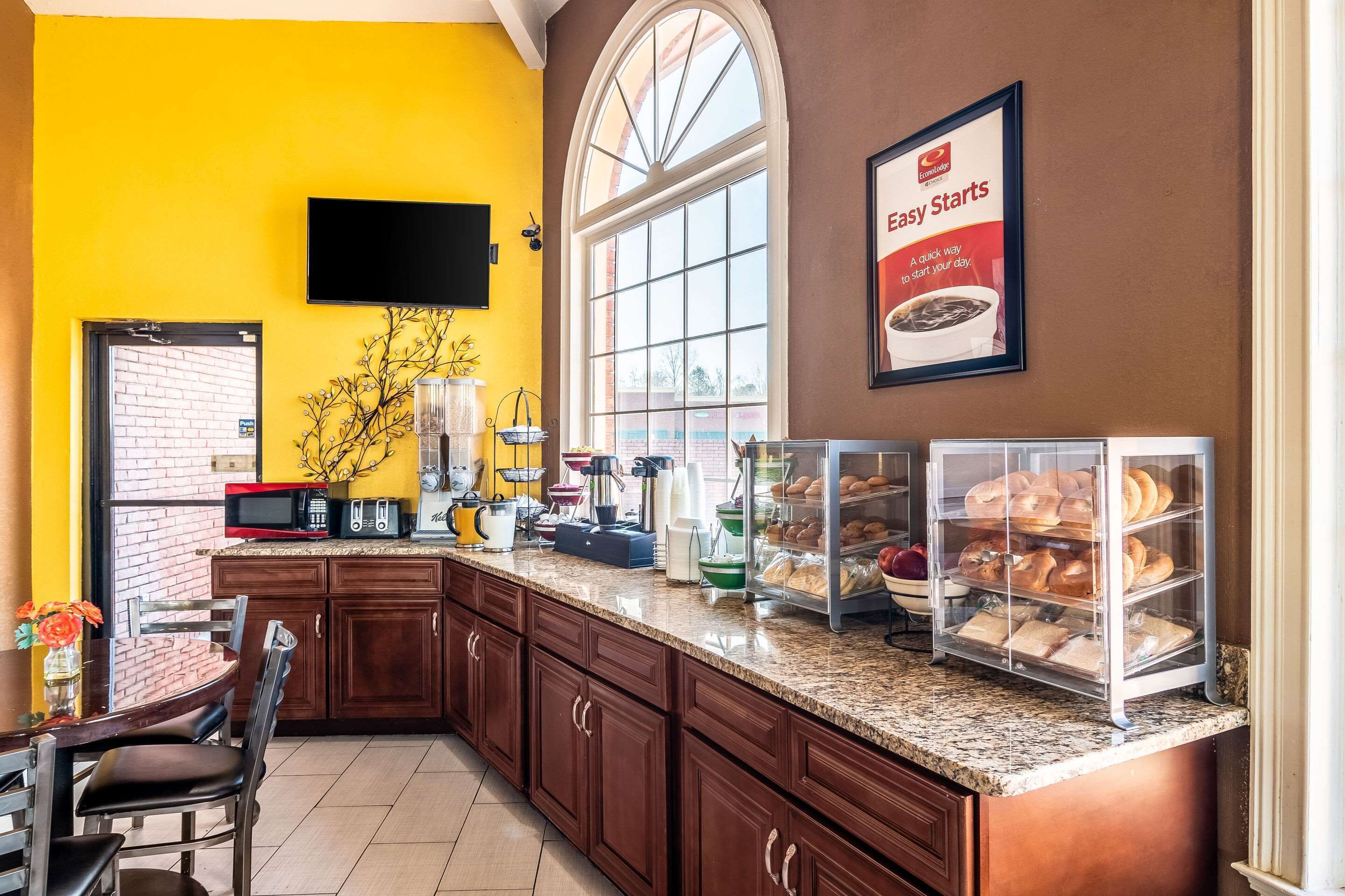A kitchen with a yellow wall, a large window, a flat-screen TV, and a bread display.