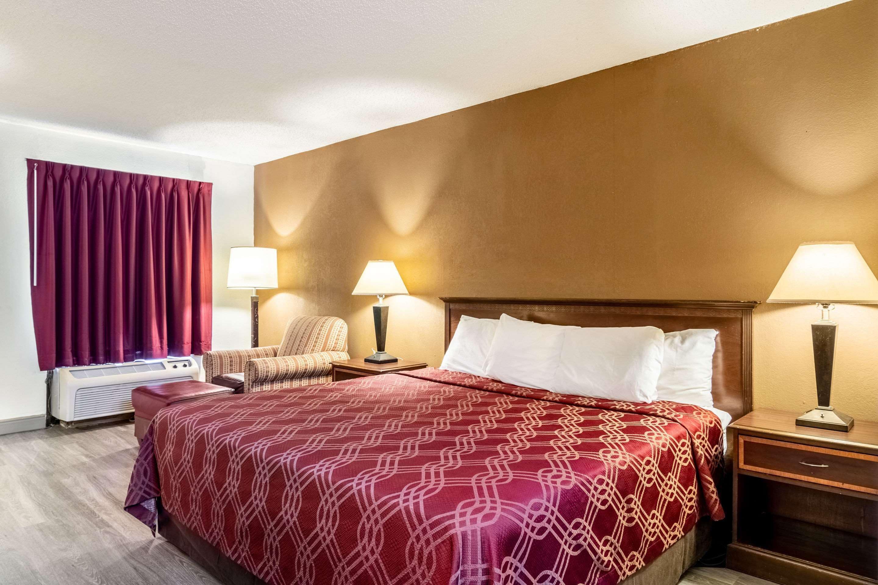 A hotel room with a large bed, two bedside tables with lamps, a chair, a window with curtains, and an air conditioning unit.