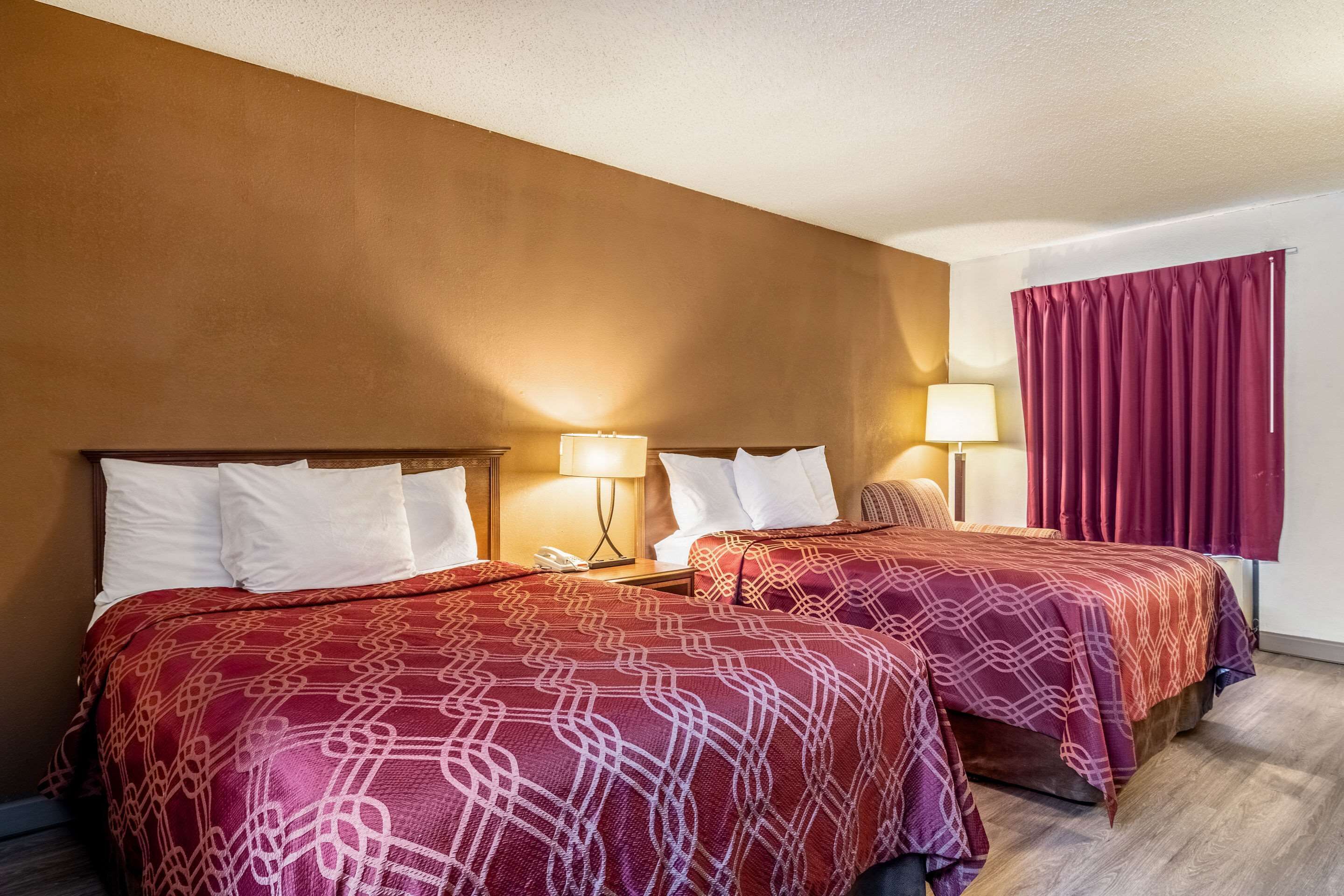 Quarto de hotel com duas camas, cada uma com roupa de cama estampada em branco e vermelho, cabeceira de madeira, abajures e cortina vermelha.